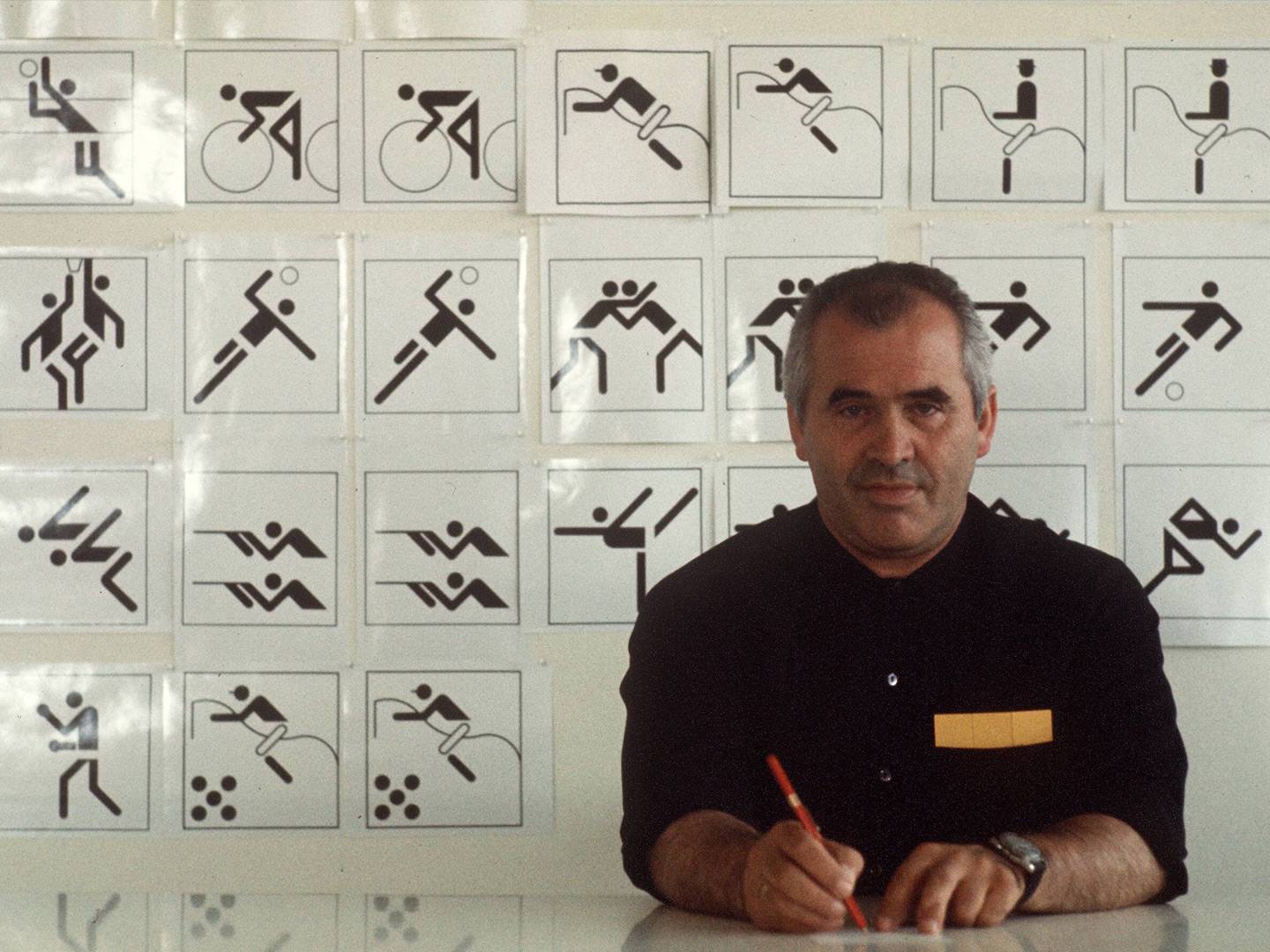 Otl Aicher sitzt vor Wand mit ikonischen Sportpiktogrammen. Sinnbild für klares Kommunikationsdesign, Markenentwicklung und visuelle Identität.